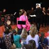 A person wearing a bright pink suit sits in the middle of a room with an audience of children sitting on the floor in front of him and a group of musicians standing behind him. 