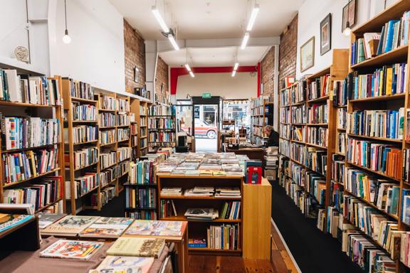 Inside The Ferret Bookshop, a book store on Cuba Street in Te Aro, Wellington. The narrow space is lined with shelves full of books and a worker sits behind the counter. 