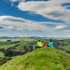 Three people sitting on a grassy hill dressed in pink, green and blue. They are looking out across the landscape of rolling hills and farmland.