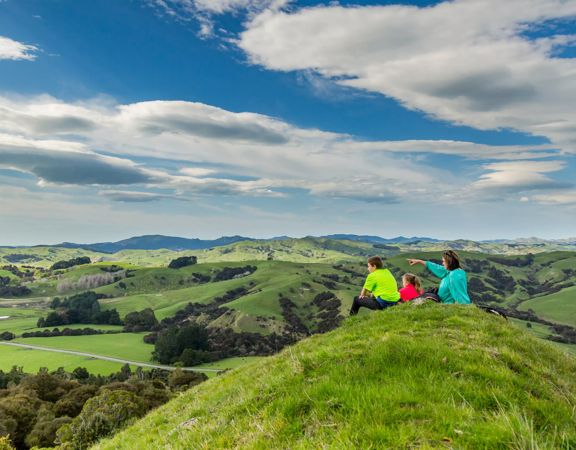 Three people sitting on a grassy hill dressed in pink, green and blue. They are looking out across the landscape of rolling hills and farmland.