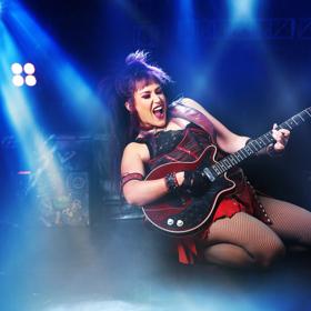 An actor performing in the We Will Rock You musical wearing a red and black leather dress, playing an electric guitar while sliding on their knees. 