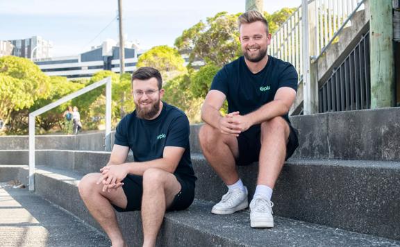 Two men wearing black tee shirts and shorts sit on cement steps on a sunny day with trees and buildings in the background.