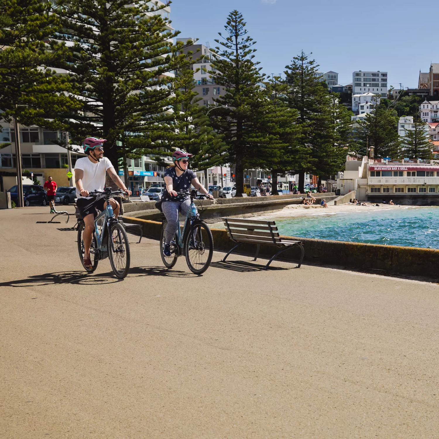 Two people ride e-bikes on the Wellington waterfront. It's a nice day and the old band rotunda is in the background.