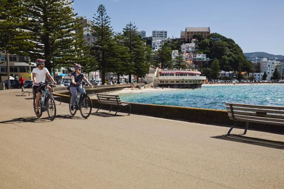 Two people ride e-bikes on the Wellington waterfront. It's a nice day and the old band rotunda is in the background.