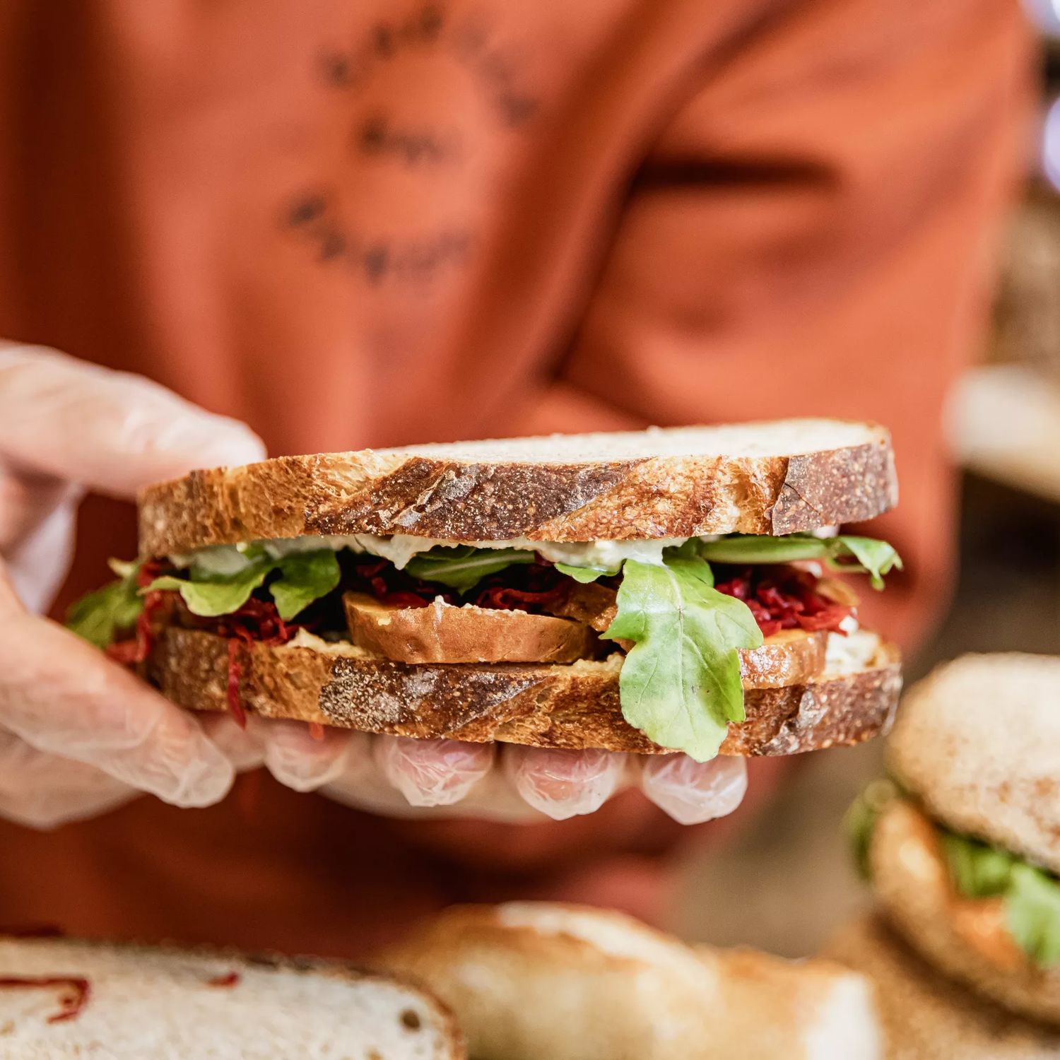 A worker wears an orange jumper and plastic gloves holds a sandwich at Shelly Bay Baker, a bakery in Te Aro, Wellington.
