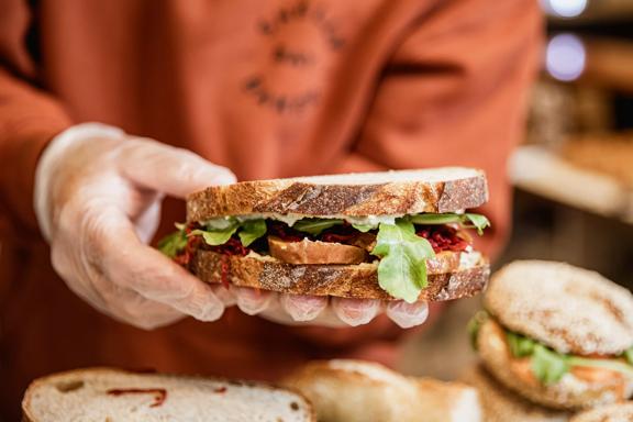 A worker wears an orange jumper and plastic gloves holds a sandwich at Shelly Bay Baker, a bakery in Te Aro, Wellington. 