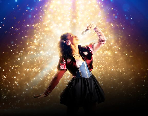 Juliet wears pink headphones, holds a microphone and sings in a sparkly spotlight.
