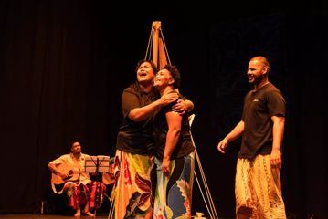 Three actors/singers are in the middle of a scene. An acoustic guitarist is in the background while a group of three are in the foreground.