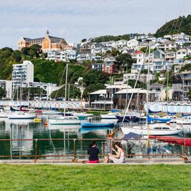 Two people sit on a curb looking at a marina and houses on a hillside. 