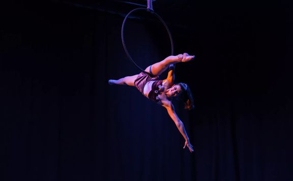 Aerialist balancing from a large suspended ring. They are holding on with one hand and doing the splits.
