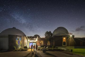 Three people stand at the front entrance of Space Place at Carter Observatory, located in Wellington Botanic Gardens on a starry clear night.