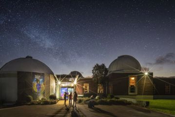 Three people stand at the front entrance of Space Place at Carter Observatory, located in Wellington Botanic Gardens on a starry clear night.