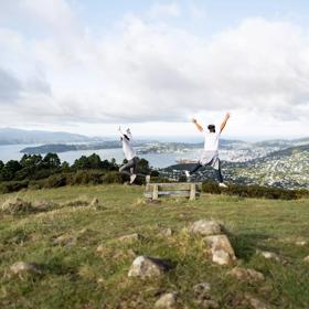 Two people jump from a park bench with outstretched arms on a grassy hill with a view of Wellington Harbour in the distance. 