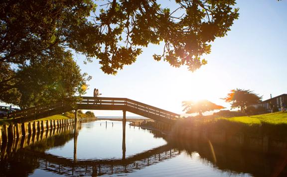 Two people walk across a small wooden bridge over a river at Raumati Beach in the Kāpiti Coast.