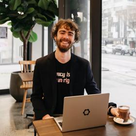 Leon Bowie looks up smiling while sitting in a café at a small table by the front window using his laptop.