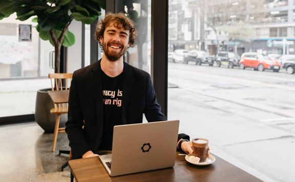 Leon Bowie looks up smiling while sitting in a café at a small table by the front window using his laptop.