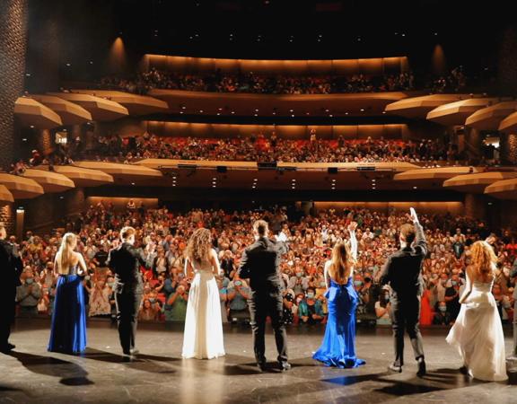 Eleven performers from World of Musicals stand in a row across centre-stage accepting a standing ovation from a full audience. 
