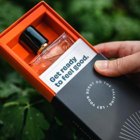 A close up shot of Speak Scents' perfume with blurry greenery in the background. The product is in it's orange and black cardboard rectangular box packaging with "Get ready to feel good." written on it.