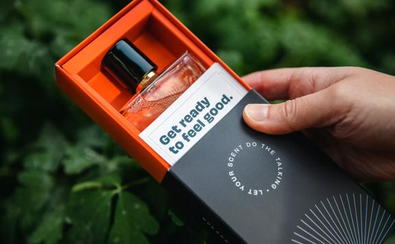 A close up shot of Speak Scents' perfume with blurry greenery in the background. The product is in it's orange and black cardboard rectangular box packaging with "Get ready to feel good." written on it.