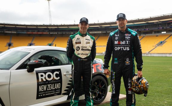 Two race drivers stand beside a white race car on the field of a large stadium.