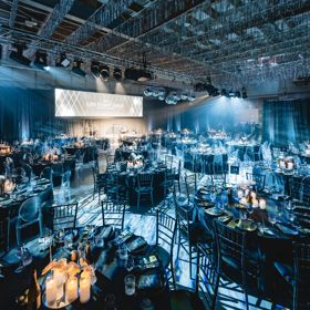 A gala set up in a flight hangar has many round tables with chairs, and dinner setting.