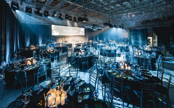 A gala set up in a flight hangar has many round tables with chairs, and dinner setting.
