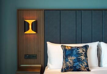 A bed at The Sebel, a 4-star hotel in Lower Hutt. The wall is dark teal, the headboard is leather and wood panel on the side with an amber lamp affixed to it and there is a blue and gold decorative pillow.