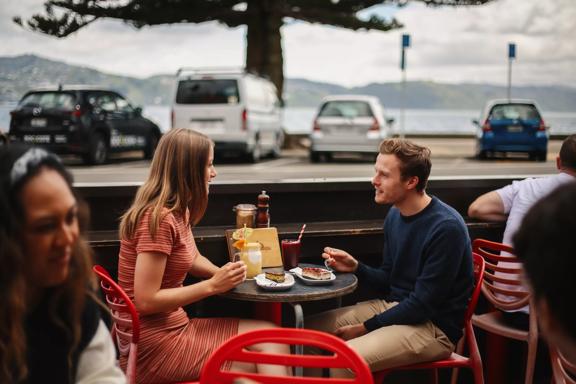2 people enjoying food and drinks at an outside table at Beach Babylon, across the road from Oriental bay.