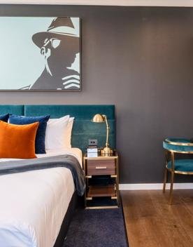 A Queen suite at Tryp by Wyndham has a bed with crisp white sheets and colour cushions against a velvet headboard. A kitchenette and table and chairs are off to the side.
