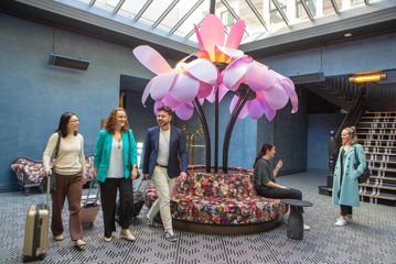 Three tourists in business casual wear walk with rolling carry-on luggage while two conference attendees are chatting inside the lobby at Naumi Hotel in Wellington. A round floral bench with a big statue of pink flowers is centred in the sunlit room.
