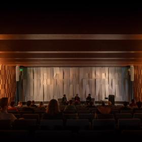 Tiakiwai conference centre auditorium view showing rows of seating and a stage with three people on chairs.