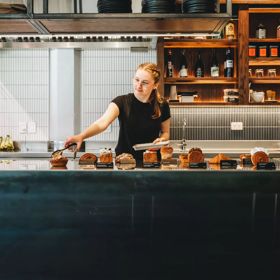 A person with tongs goes to pick up a pastry from the counter in Glou Glou. There is a row of decadent pastries.