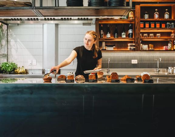 A person with tongs goes to pick up a pastry from the counter in Glou Glou. There is a row of decadent pastries.