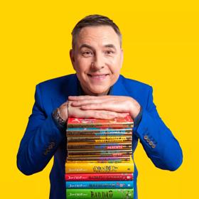 Comedian David Walliams stands with a pile of his children's books, his hands resting on the top. He wears a blue suit.