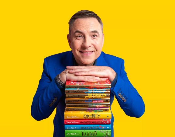 Comedian David Walliams stands with a pile of his children's books, his hands resting on the top. He wears a blue suit.
