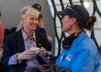 A person with short grey hair, wearing a purple patterned collared shirt, a black blazer, and an LGBT flag pin on their left lapel, smiles and holds a cappuccino with both hands while talking to a woman in a navy blue baseball cap and blue Wellington Zoo 