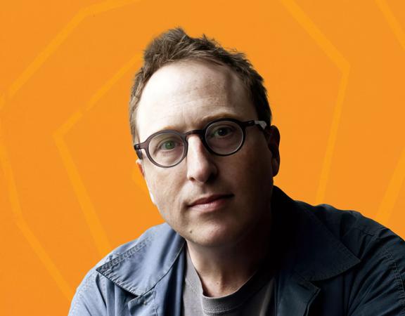 Jon Ronson, a British-American journalist, author, and filmmaker, in front of an orange background.
