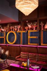 Inside the lobby of The Intrepid Hotel, where a neon sign on the wall reads HOTEL and other pink neon lights surround maroon furnishings.