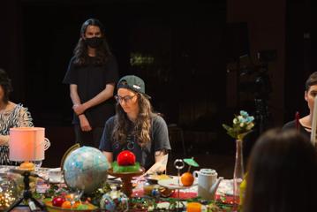 Casey Zilbert sits between two other people at a table covered in eclectic items and a person is wearing all black and a face mask standing behind them in the background.