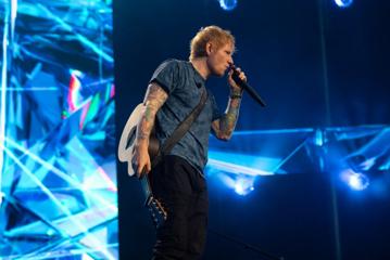 Ed Sheeran sings into a microphone with blue stage lights behind him.