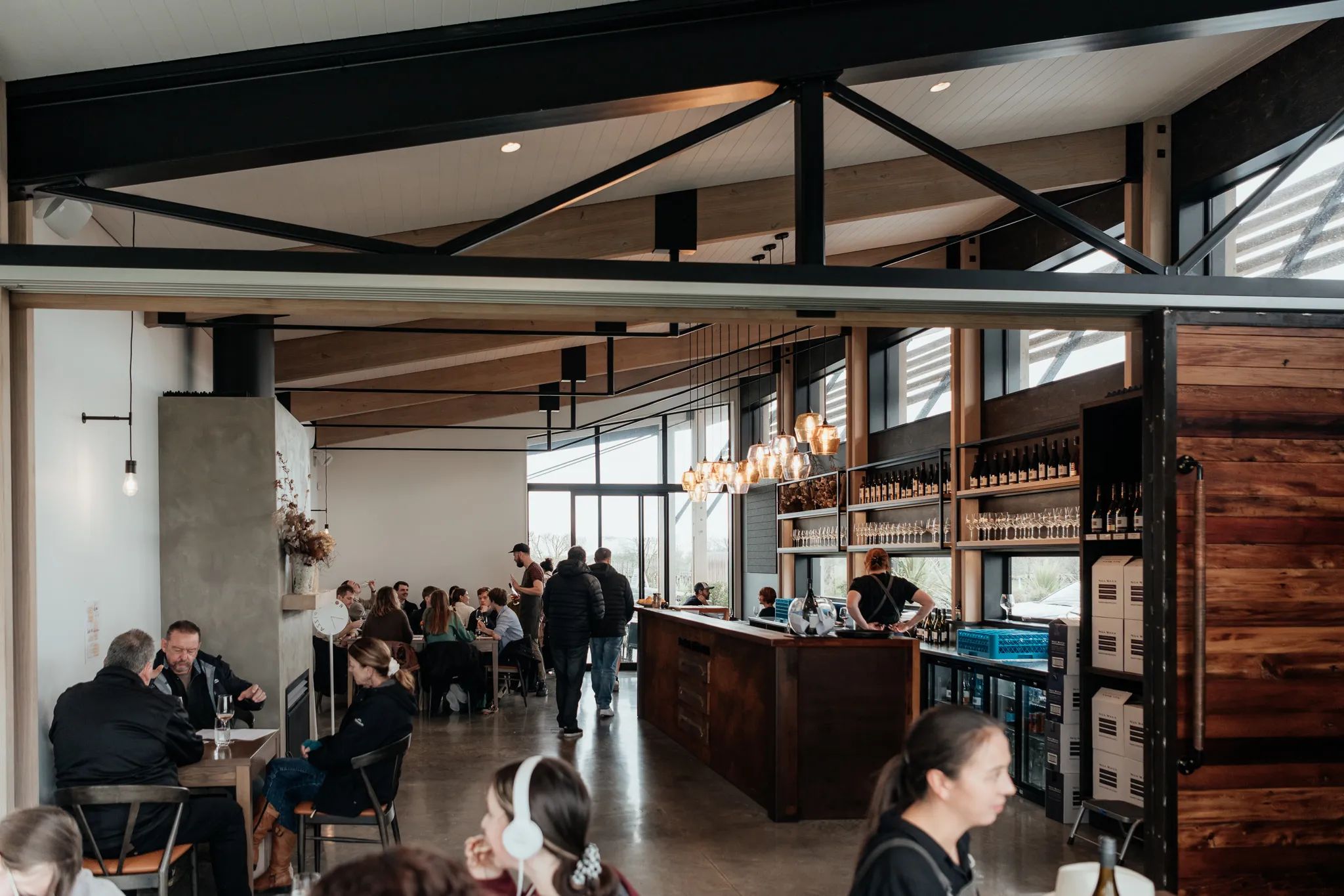The interior of Nga Waka Wines in the Wairarapa region. High wooden ceilings with black industrial poles frame the space, featuring polished concrete floors and warm lighting. People sit at tables enjoying food and drink.