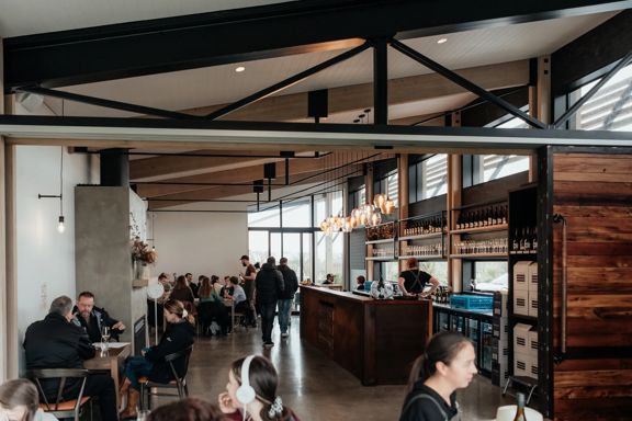 The interior of Nga Waka Wines in the Wairarapa region. High wooden ceilings with black industrial poles frame the space, featuring polished concrete floors and warm lighting. People sit at tables enjoying food and drink.