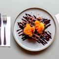A plate with waffles, chocolate sauce and oranges on a table with a fork and knife on the right and a lunch menu on the left. 