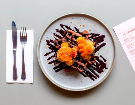 A plate with waffles, chocolate sauce and oranges on a table with a fork and knife on the right and a lunch menu on the left. 