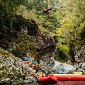 A person wearing a blue helmet, orange lifejacket, is in mid-air, doing a cannonball jump from a tall rock into a stream. Three people watch from below, where they wait with two small red rafts.