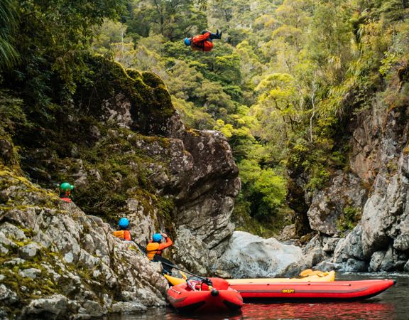 A person wearing a blue helmet, orange lifejacket, is in mid-air, doing a cannonball jump from a tall rock into a stream. Three people watch from below, where they wait with two small red rafts.