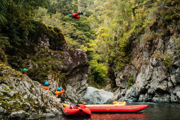 A person wearing a blue helmet, orange lifejacket, is in mid-air, doing a cannonball jump from a tall rock into a stream. Three people watch from below, where they wait with two small red rafts.