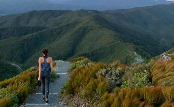 A person walks down the gravel path on the Te Ara Tirohanga track with views of the hills. The sky is tinged pink.