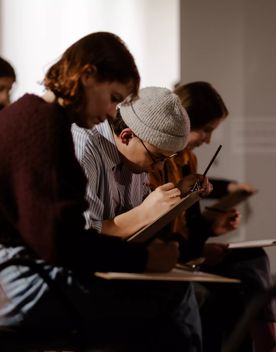 A young woman with short brown hair wearing a dark jumper, and a man wearing a grey knit hat sit and draw in a gallery space.
