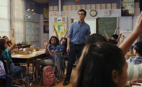 A still from the Mr. Corman TV show. Joseph Gordon-Levitt as Mr Corman stands in the middle of a classroom surrounded by children sitting at desks with their hands raised.